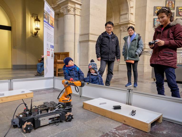 Menschen vor kleinem Fahrzeug mit Roboterarm.